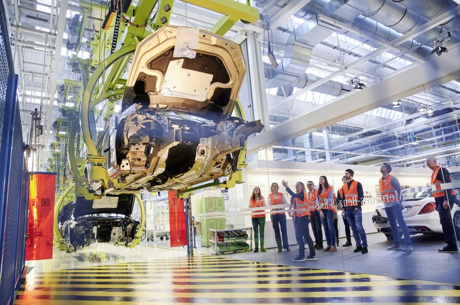 Besucher in orangefarbenen Westen beobachten die Fahrzeugmontage in einer Mercedes-Benz Produktionshalle in Sindelfingen., © Mercedes-Benz AG