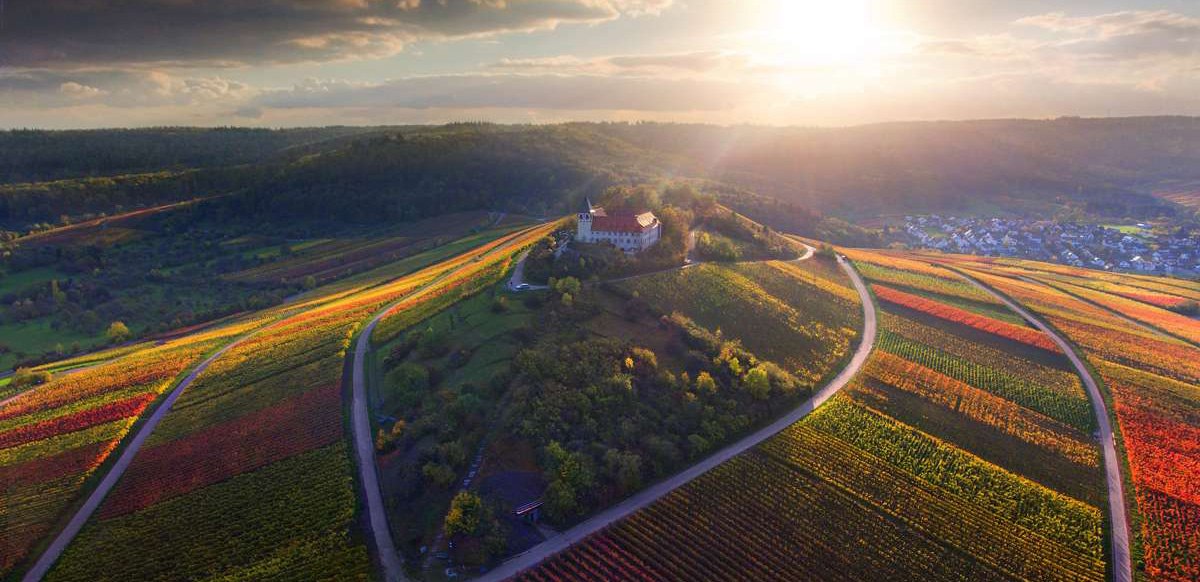 Luftaufnahme des Michaelsbergs, umgeben von bunten Weinbergen im Sonnenuntergang. Eine Kirche steht auf dem Hügel, umgeben von Wald und Feldern., © Neckar-Zaber-Tourismus e.V