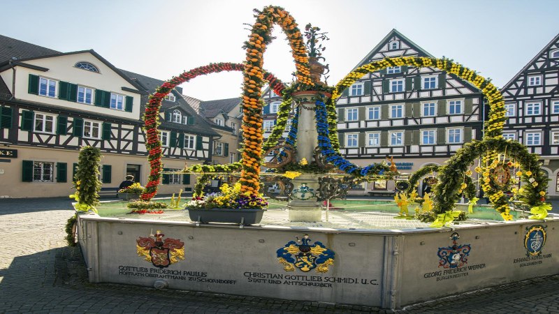 Osterbrunnen auf dem Schorndorfer Marktplatz, geschmückt mit bunten Blumen und Girlanden, umgeben von Fachwerkhäusern., © SMG, Sarah Schmid