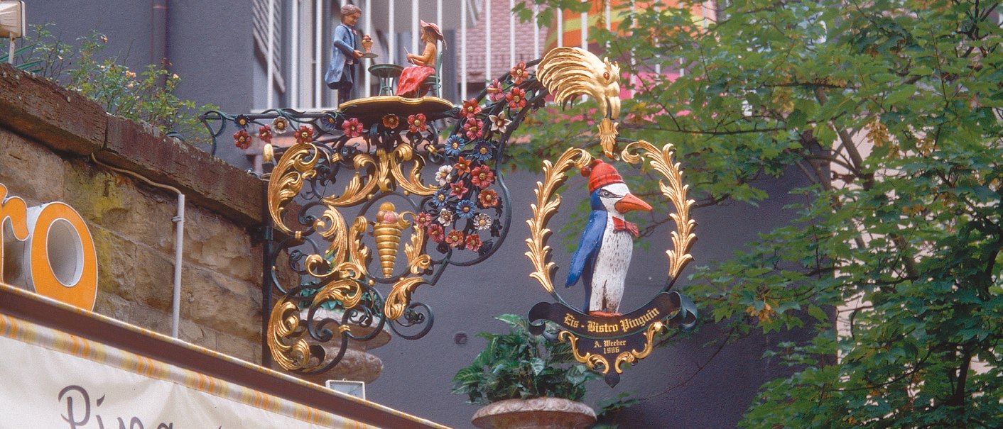 Ein kunstvoll verziertes Schild mit einem Pinguin und Figuren, umgeben von Blumenornamenten, hängt an einer Wand am Eugensplatz., © Stuttgart-Marketing GmbH Ein kunstvoll verziertes Schild mit einem Pinguin und Figuren, umgeben von Blumenornamenten, hängt an einer Wand am Eugensplatz., © Stuttgart-Marketing GmbH