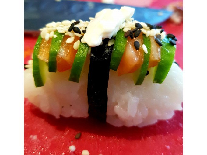 Sushi-Nigiri mit Lachs, Gurke, Sesam und Wasabi auf rotem Untergrund. Ein Streifen Nori h&auml;lt die Zutaten zusammen., &copy; Kultweine-Kochwelten-Kunst&uuml;cke - Kochschule Sonja Lenz