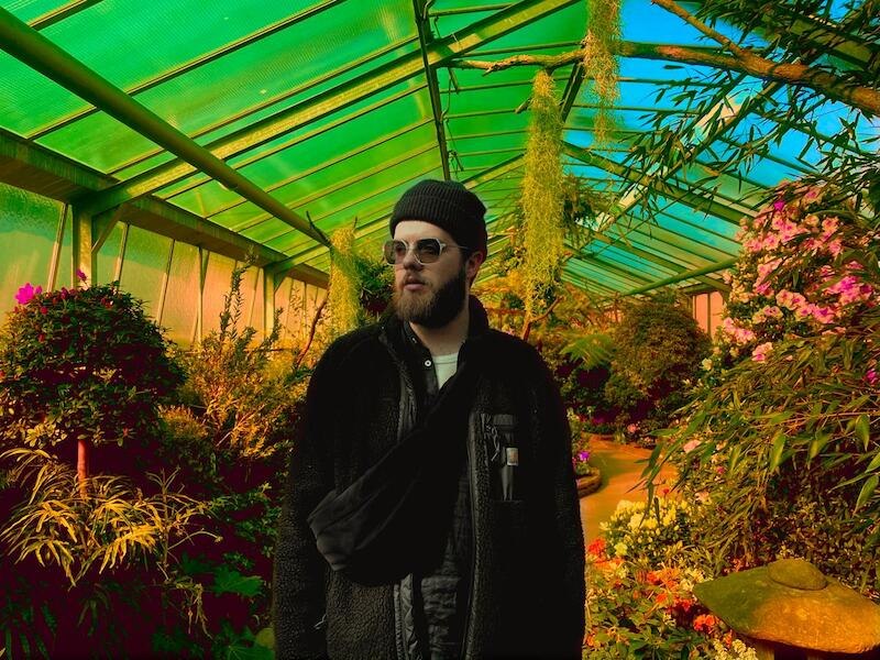 Eine Person mit Sonnenbrille und M&uuml;tze steht in einem farbenfrohen Gew&auml;chshaus voller Pflanzen und Blumen., &copy; Interessengemeinschaft Kultur Sindelfingen / B&ouml;blingen e.V.