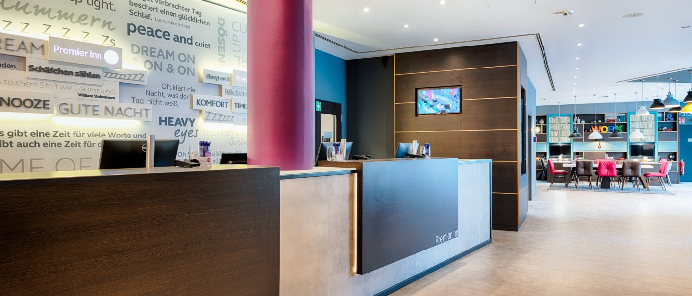 Moderne Hotelrezeption von Premier Inn mit Schriftzügen an der Wand und einem angrenzenden Sitzbereich mit Tischen und Stühlen., © Premier Inn Germany Moderne Hotelrezeption von Premier Inn mit Schriftzügen an der Wand und einem angrenzenden Sitzbereich mit Tischen und Stühlen., © Premier Inn Germany
