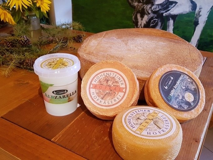 Verschiedene Käsesorten und ein Becher Albzarella auf einem Holztisch, dekoriert mit Sonnenblumen und Tannenzapfen., © SwabianTravel Verschiedene Käsesorten und ein Becher Albzarella auf einem Holztisch, dekoriert mit Sonnenblumen und Tannenzapfen., © SwabianTravel