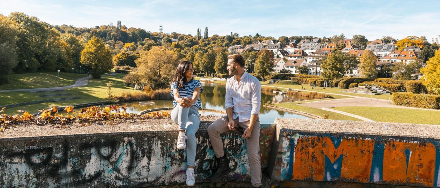 Zwei Personen sitzen auf einer bemalten Mauer im Wartbergpark. Im Hintergrund sind ein Teich, Bäume und Häuser zu sehen. Es ist ein sonniger Tag., © Stuttgart-Marketing GmbH, Sarah Schmid
