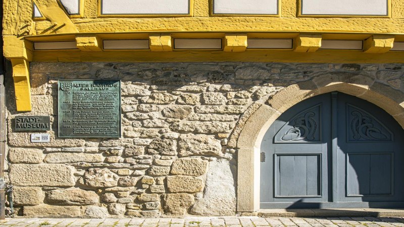 Nahaufnahme des Stadtmuseums Sindelfingen mit gelbem Fachwerk, blauer Tür und Infotafel an einer Steinwand., © SMG,  Sarah Schmid Nahaufnahme des Stadtmuseums Sindelfingen mit gelbem Fachwerk, blauer Tür und Infotafel an einer Steinwand., © SMG,  Sarah Schmid