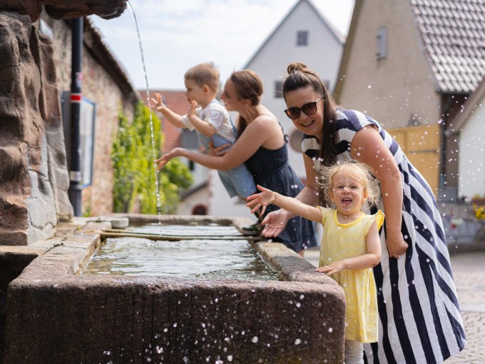 Zwei Frauen und zwei Kinder spielen fr&ouml;hlich an einem Brunnen in einem malerischen Dorf. Die Kinder planschen im Wasser, w&auml;hrend die Frauen l&auml;cheln., &copy; Stadtverwaltung Weil der Stadt