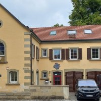 zweist&ouml;ckiges historisches Geb&auml;ude mit gelber Fassade und roten Dachziegeln. Im Untergescho&szlig; auf der rechten Seite drei rotbraune Doppelt&uuml;ren. Vor den T&uuml;ren stehen ein schwarzer PKW und zweiAnh&auml;nger, &copy; Petra Natzkowski