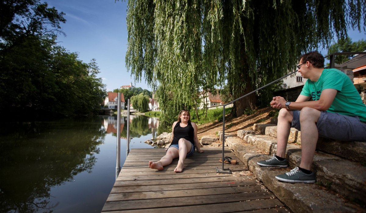 Zwei Personen sitzen entspannt auf einem Holzsteg am Flussufer. Eine Frau sitzt barfuß, ein Mann sitzt auf Steinstufen. Im Hintergrund sind Bäume und Häuser., © Stoppel