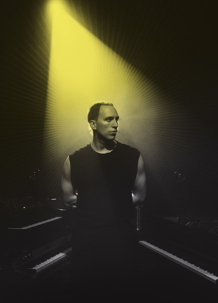 Ein Mann in schwarzem Shirt steht im gelben Scheinwerferlicht zwischen zwei Keyboards, umgeben von Dunkelheit., © Scala Kultur Live gGmbH Ein Mann in schwarzem Shirt steht im gelben Scheinwerferlicht zwischen zwei Keyboards, umgeben von Dunkelheit., © Scala Kultur Live gGmbH