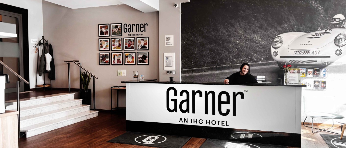 Moderne Hotelrezeption mit einem Empfangstresen, an dem eine Mitarbeiterin l&auml;chelt. An der Wand ein gro&szlig;es Foto eines Rennwagens., &copy; Novum Hospitality