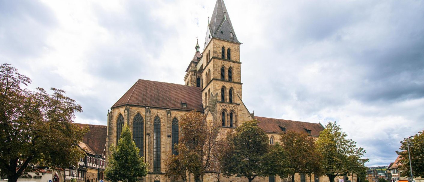 Die Stadtkirche St. Dionys in Esslingen mit ihrem markanten Turm, umgeben von Bäumen und historischen Gebäuden, unter einem bewölkten Himmel., © Stuttgart-Marketing GmbH, Sarah Schmid