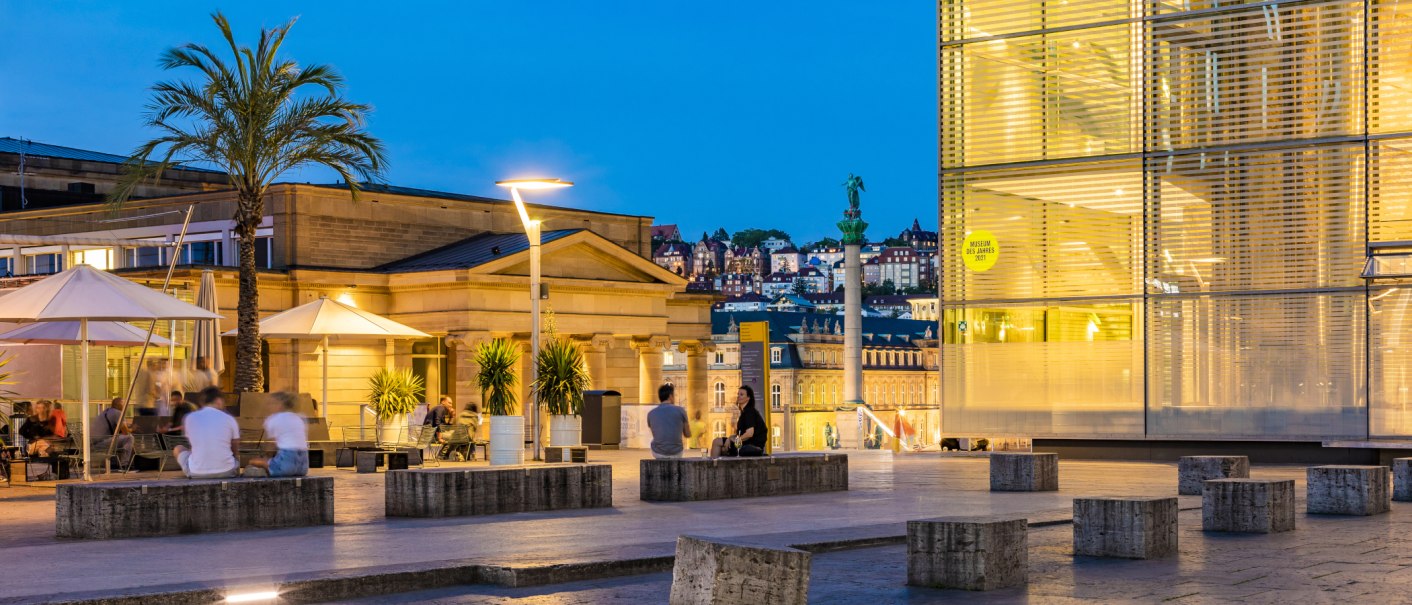 Abendstimmung am Kleinen Schlossplatz mit Palmen, Menschen und moderner Architektur. Im Hintergrund sind beleuchtete Gebäude und eine Statue zu sehen., © SMG, Werner Dieterich Abendstimmung am Kleinen Schlossplatz mit Palmen, Menschen und moderner Architektur. Im Hintergrund sind beleuchtete Gebäude und eine Statue zu sehen., © SMG, Werner Dieterich