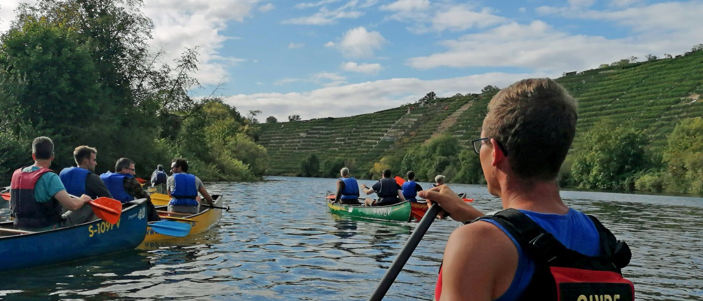Eine Gruppe von Menschen paddelt in Kanus auf einem Fluss, umgeben von grünen Hügeln. Ein Guide ist im Vordergrund zu sehen., © Die Zugvögel
