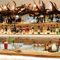 Eine Bar mit einer Holztheke, auf der verschiedene Getränke stehen. Im Hintergrund sind Regale mit Spirituosen und eine Geweih-Lampe zu sehen., © TOMAS Eine Bar mit einer Holztheke, auf der verschiedene Getränke stehen. Im Hintergrund sind Regale mit Spirituosen und eine Geweih-Lampe zu sehen., © TOMAS