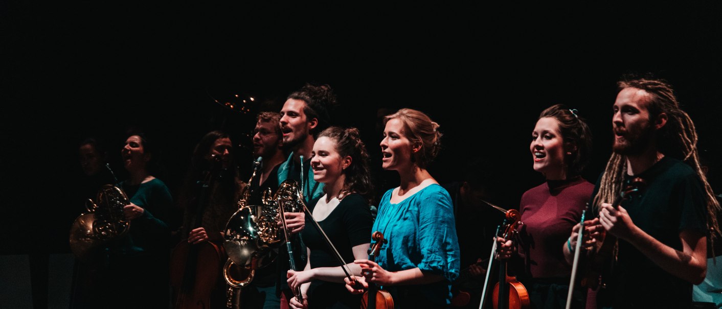 Musiker auf einer Bühne mit Hörnern, Geigen und Flöten, in dunkler Umgebung, lächelnd und konzentriert., © Sky Bürhaus Musiker auf einer Bühne mit Hörnern, Geigen und Flöten, in dunkler Umgebung, lächelnd und konzentriert., © Sky Bürhaus