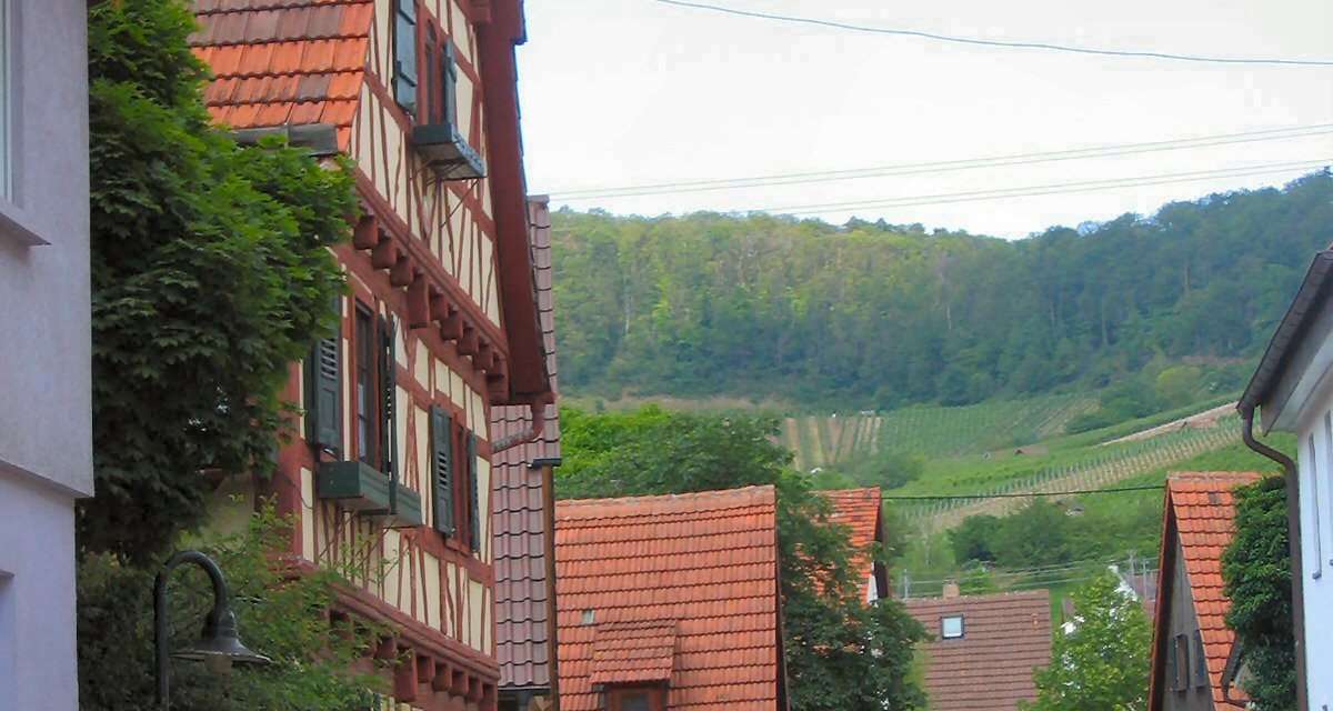 Fachwerkhäuser säumen eine ruhige Dorfstraße. Im Hintergrund sind grüne Weinberge zu sehen. Autos parken am Straßenrand., © Land der 1000 Hügel - Kraichgau-Stromberg Fachwerkhäuser säumen eine ruhige Dorfstraße. Im Hintergrund sind grüne Weinberge zu sehen. Autos parken am Straßenrand., © Land der 1000 Hügel - Kraichgau-Stromberg