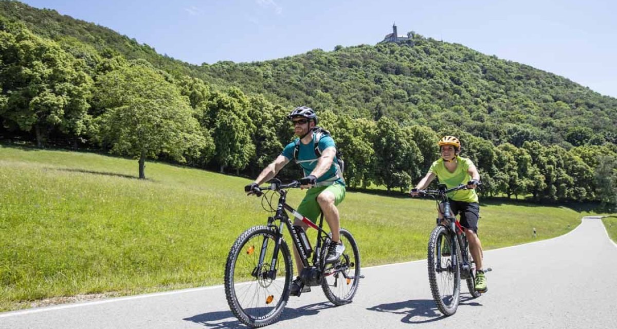 Zwei Mountainbiker fahren auf einer Stra&szlig;e durch eine gr&uuml;ne Landschaft. Im Hintergrund ist ein bewaldeter H&uuml;gel mit einem Turm zu sehen., &copy; Schw&auml;bische Alb Tourismusverband e.V.
