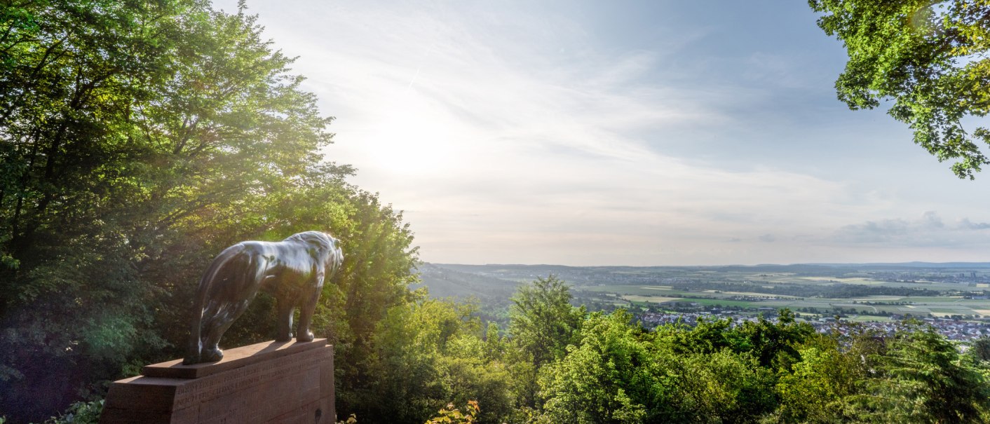 Eine Bronzestatue eines Löwen steht auf einem Sockel, umgeben von Bäumen, mit Blick auf eine weite Landschaft unter einem klaren Himmel., © SMG, Martina Denker Eine Bronzestatue eines Löwen steht auf einem Sockel, umgeben von Bäumen, mit Blick auf eine weite Landschaft unter einem klaren Himmel., © SMG, Martina Denker