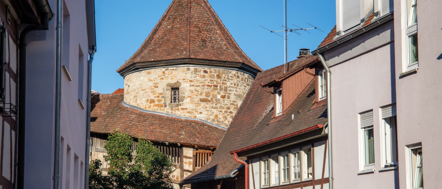 Der Blockturm in Nürtingen ist zwischen zwei Häusern sichtbar. Der Turm hat ein rundes, steinernes Mauerwerk und ein spitzes, rotes Ziegeldach., © SMG, Achim Mende