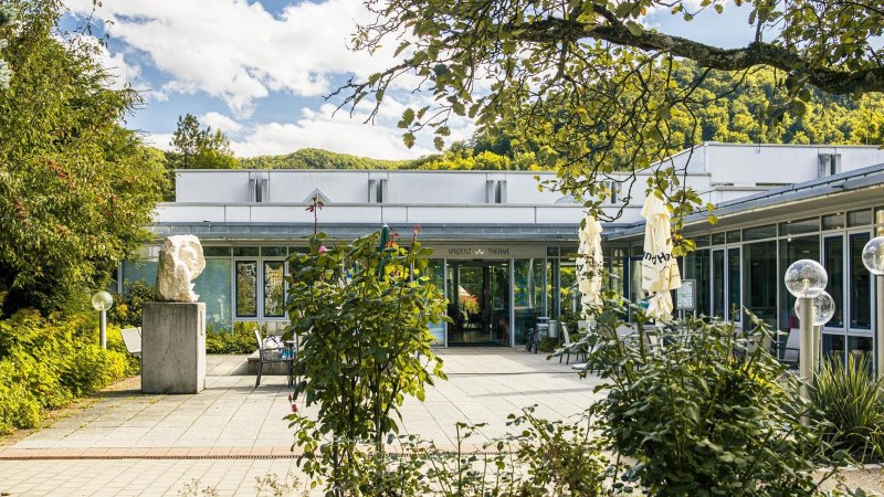 Eingang der Vinzenz Therme in Bad Ditzenbach, umgeben von B&auml;umen und Pflanzen. Moderne Architektur mit Glasfronten und Skulpturen im Vordergrund., &copy; Stuttgart-Marketing GmbH, Sarah Schmid