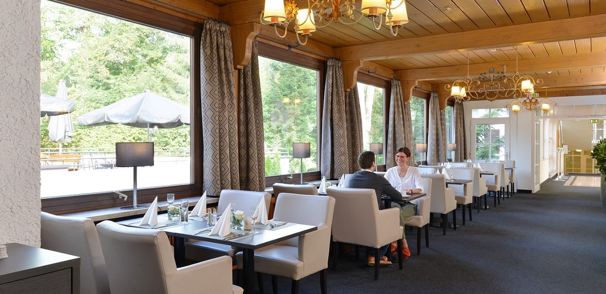 Elegantes Restaurant mit großen Fenstern, weißen Sesseln und Holzelementen. Zwei Personen sitzen an einem Tisch. Draußen sind Sonnenschirme sichtbar., © relexa Waldhotel Schatten Elegantes Restaurant mit großen Fenstern, weißen Sesseln und Holzelementen. Zwei Personen sitzen an einem Tisch. Draußen sind Sonnenschirme sichtbar., © relexa Waldhotel Schatten
