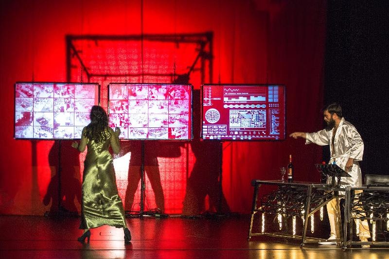 Zwei Personen auf einer Bühne mit rotem Hintergrund. Eine Frau in einem goldenen Kleid steht vor Bildschirmen, ein Mann in einem weißen Mantel zeigt auf etwas., © Backnang