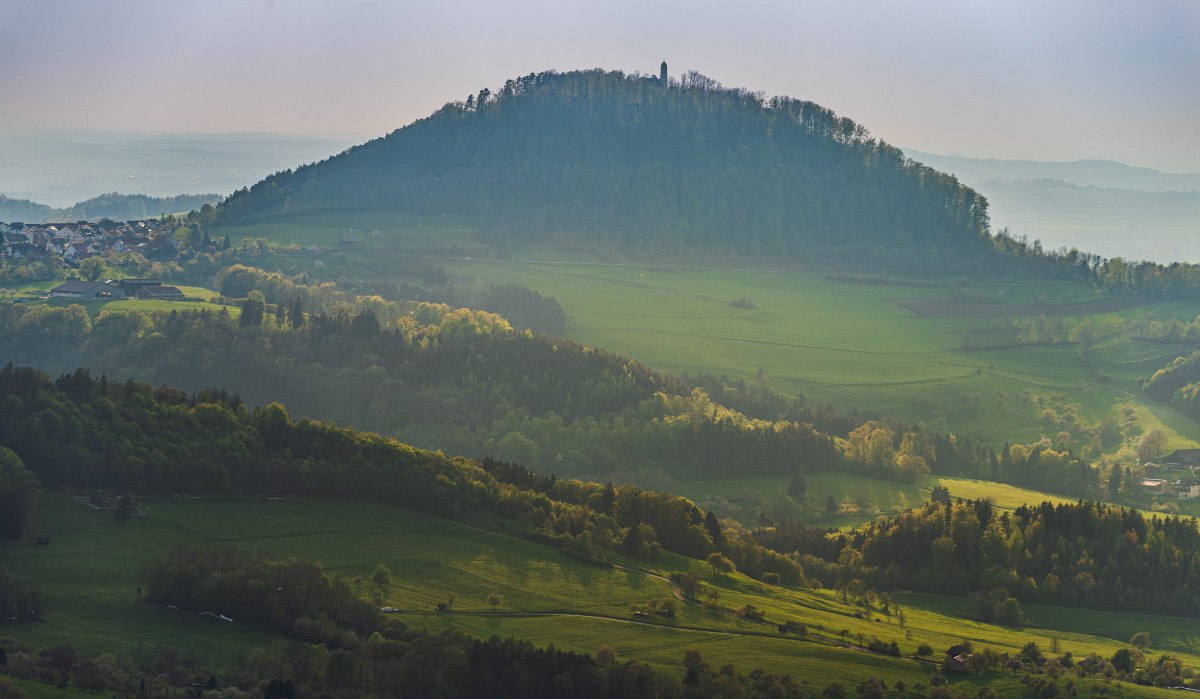Der Rechberg erhebt sich majest&auml;tisch &uuml;ber die umliegenden gr&uuml;nen Wiesen und W&auml;lder, w&auml;hrend die Sonne sanft &uuml;ber die Landschaft scheint.