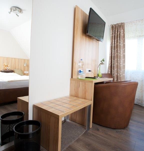 Hotelzimmer mit Schreibtisch, Stuhl, Wand-TV und Bett im Hintergrund. Helle Einrichtung mit Holzdetails und Vorhängen., © Hotel Reinhardtshof Garni