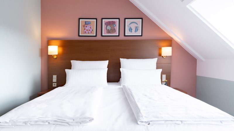 Ein stilvoll eingerichtetes Hotelzimmer mit einem gro&szlig;en Doppelbett, wei&szlig;en Bettlaken und Kissen. Drei Bilder schm&uuml;cken die Wand &uuml;ber dem Bett., &copy; Novum  Hospitality