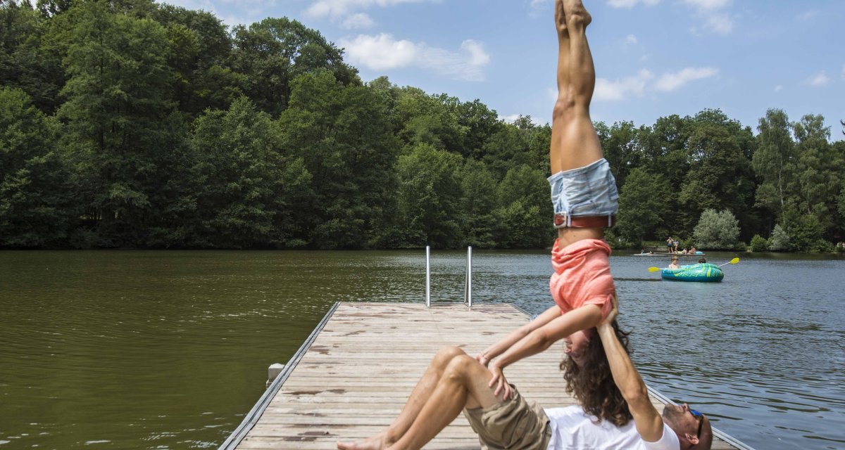 Ein Paar macht eine Akrobatik&uuml;bung auf einem Steg an einem See, umgeben von Wald. Im Hintergrund ist ein aufblasbares Boot zu sehen., &copy; Schw&auml;bische Alb Tourismusverband e.V.