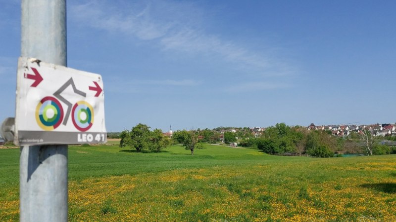 Ein Fahrradweg-Schild an einem Pfosten vor einer gr&uuml;nen Wiese mit gelben Blumen. Im Hintergrund ist ein Dorf zu sehen., &copy; Natur.Nah. Sch&ouml;nbuch & Heckeng&auml;u