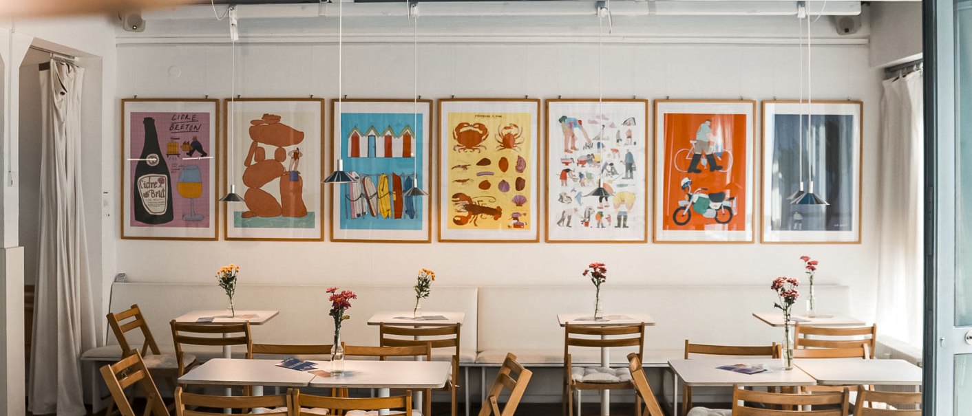Ein gemütliches Café mit Holzmöbeln, bunten Postern an der Wand und Blumen auf den Tischen. Ein Projektor hängt an der Decke., © Merlin, Stuttgart Ein gemütliches Café mit Holzmöbeln, bunten Postern an der Wand und Blumen auf den Tischen. Ein Projektor hängt an der Decke., © Merlin, Stuttgart