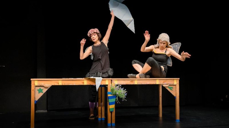Zwei Schauspielerinnen stehen auf einem Tisch. Eine trägt Flügel, die andere hält einen Papierdrachen. Beide sind in einer Theaterszene vertieft., © Katrin Schander Zwei Schauspielerinnen stehen auf einem Tisch. Eine trägt Flügel, die andere hält einen Papierdrachen. Beide sind in einer Theaterszene vertieft., © Katrin Schander