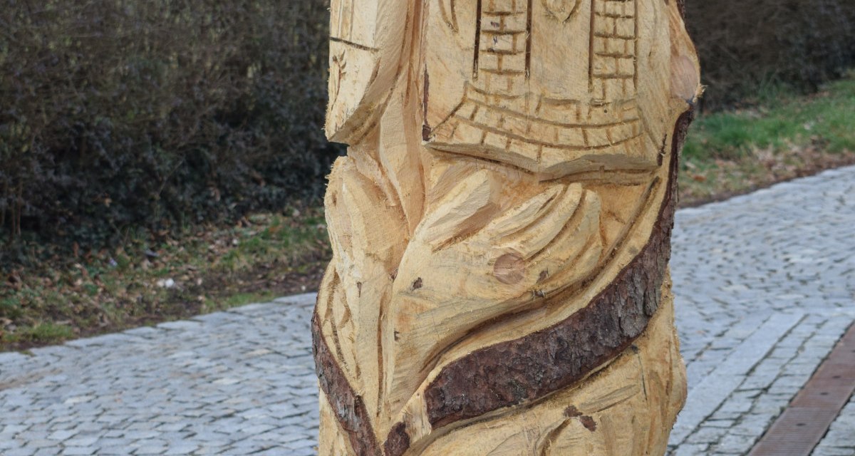 Holzskulptur mit geschnitzten Motiven, darunter ein Gesicht und Gebäude, auf einem gepflasterten Weg am Skulpturenpfad Böbingen., © Gemeinde Böbingen Holzskulptur mit geschnitzten Motiven, darunter ein Gesicht und Gebäude, auf einem gepflasterten Weg am Skulpturenpfad Böbingen., © Gemeinde Böbingen