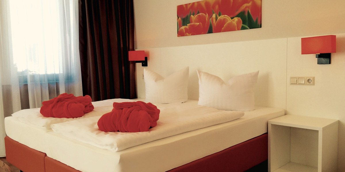 Ein gemütliches Hotelzimmer mit Doppelbett, roten Bademänteln und einem Tulpenbild an der Wand. Die Beleuchtung ist warm und einladend., © TOMAS