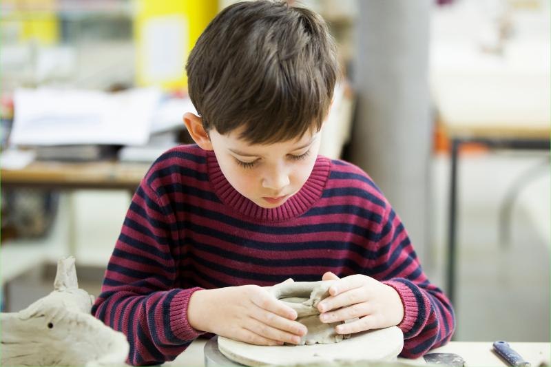 Ein Junge in einem gestreiften Pullover formt konzentriert Ton in einem Workshop. Im Hintergrund sind unscharfe Tische und Materialien zu sehen., © Kunstschule Labyrinth Ein Junge in einem gestreiften Pullover formt konzentriert Ton in einem Workshop. Im Hintergrund sind unscharfe Tische und Materialien zu sehen., © Kunstschule Labyrinth