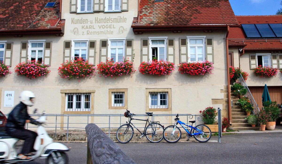 Ein traditionelles Gebäude mit roten Dachziegeln und Blumenkästen. Ein Rollerfahrer fährt vorbei, zwei Fahrräder stehen vor dem Haus., © Waiblingen - Stuttgart-Marketing GmbH Ein traditionelles Gebäude mit roten Dachziegeln und Blumenkästen. Ein Rollerfahrer fährt vorbei, zwei Fahrräder stehen vor dem Haus., © Waiblingen - Stuttgart-Marketing GmbH