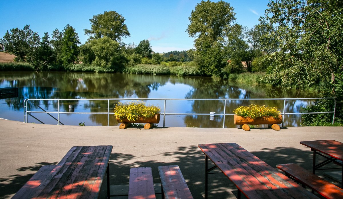 Ein idyllischer See mit Bäumen im Hintergrund, umgeben von Natur. Im Vordergrund stehen Bänke und Blumenkästen mit gelben Blumen., © agentur arcos/Niki Eilers Ein idyllischer See mit Bäumen im Hintergrund, umgeben von Natur. Im Vordergrund stehen Bänke und Blumenkästen mit gelben Blumen., © agentur arcos/Niki Eilers