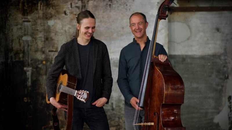 Zwei Männer stehen lächelnd mit Gitarre und Kontrabass vor einer rustikalen Wand., © Foto Rochus Enssle Zwei Männer stehen lächelnd mit Gitarre und Kontrabass vor einer rustikalen Wand., © Foto Rochus Enssle