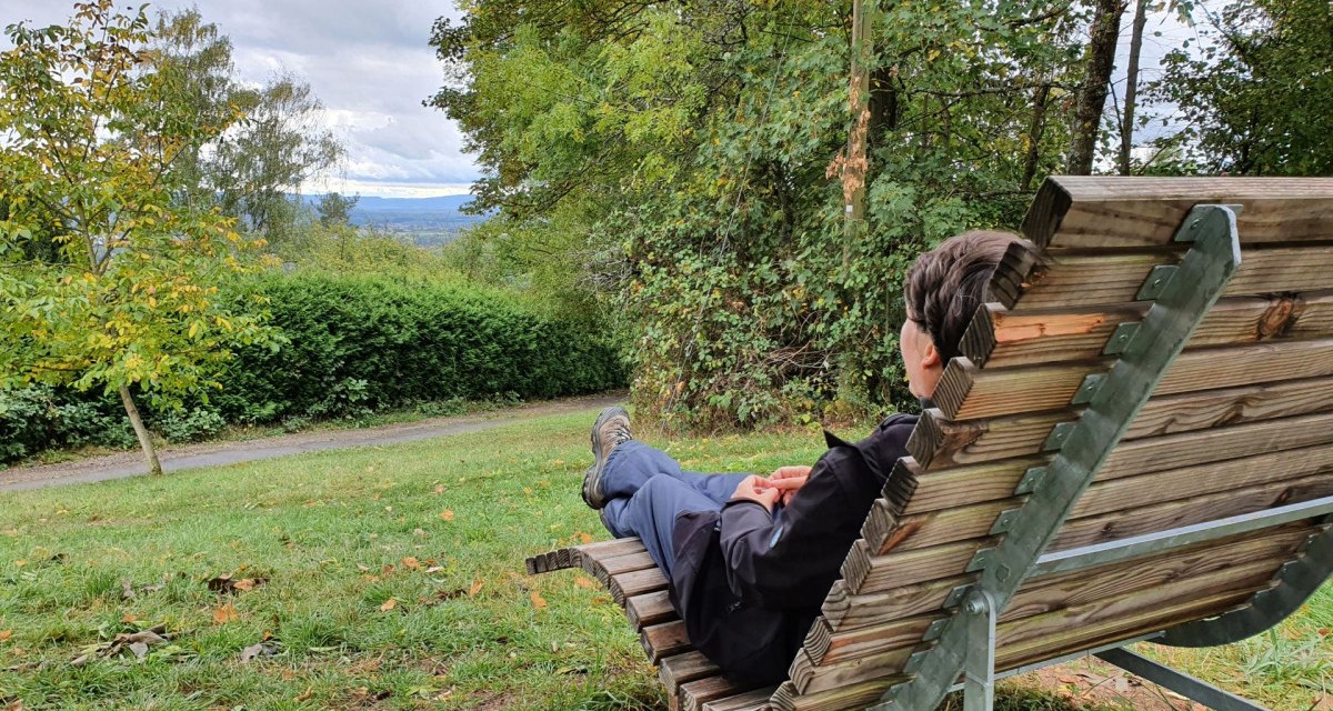 Eine Person sitzt entspannt auf einer Holzbank und blickt auf eine grüne, bewaldete Landschaft mit bewölktem Himmel. Eine Person sitzt entspannt auf einer Holzbank und blickt auf eine grüne, bewaldete Landschaft mit bewölktem Himmel.