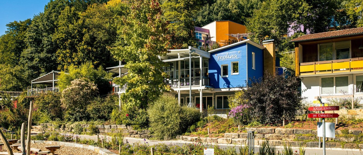 Ein blaues Geb&auml;ude namens 'molina' inmitten einer gr&uuml;nen, bewaldeten Landschaft mit bunten Pflanzen und einem Wegweiser im Vordergrund., &copy; Stuttgart-Marketing GmbH, Sarah Schmid