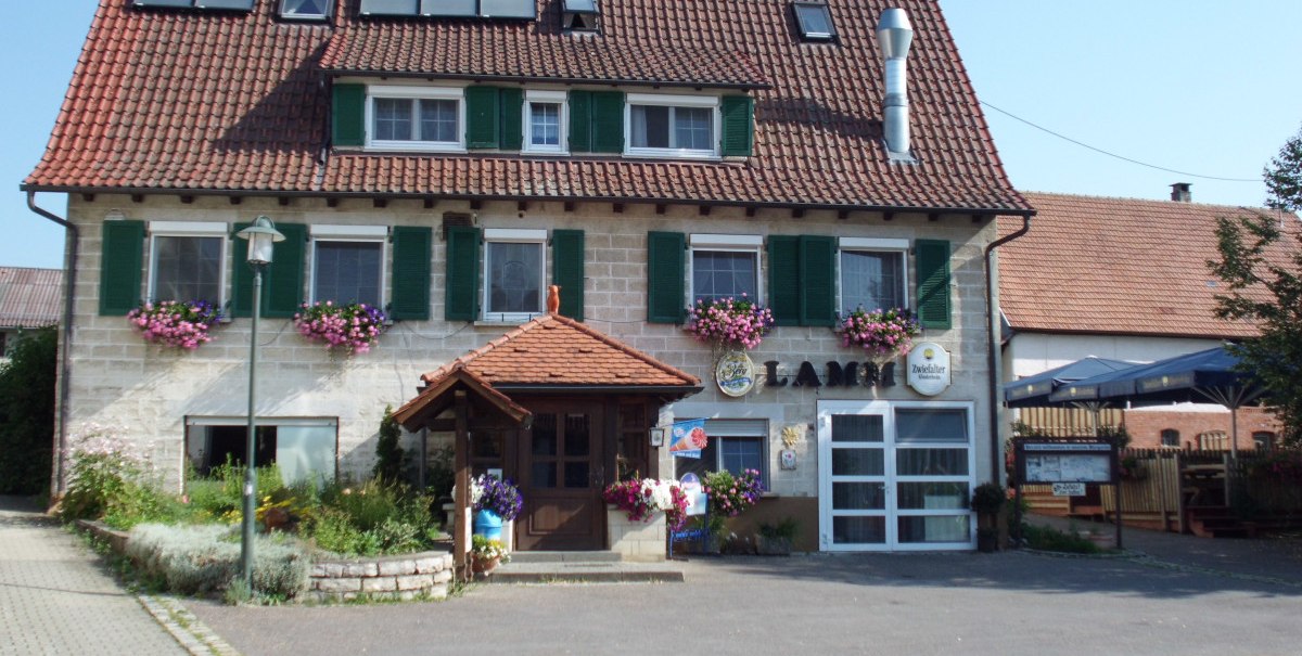 Ein traditionelles Gasthaus mit roten Dachziegeln, grünen Fensterläden und Blumenkästen. Ein Biergarten ist rechts zu sehen. Ein traditionelles Gasthaus mit roten Dachziegeln, grünen Fensterläden und Blumenkästen. Ein Biergarten ist rechts zu sehen.