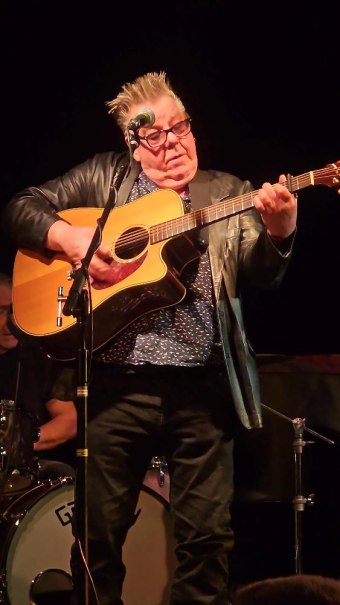 Ein Gitarrist mit Brille und Lederjacke spielt konzentriert auf der B&uuml;hne, w&auml;hrend ein Schlagzeuger im Hintergrund zu sehen ist., &copy; Foto Bernd Hoffmann