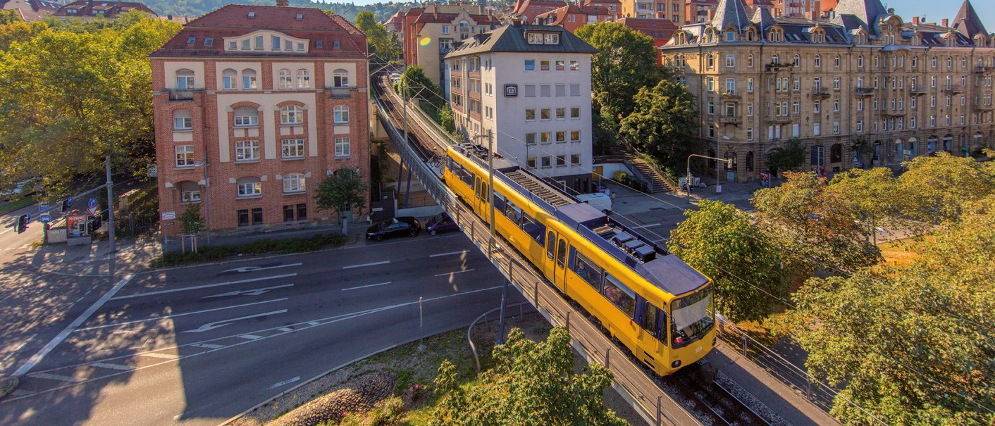 Die Zahnradbahn in Stuttgart fährt auf einer erhöhten Schiene durch eine städtische Umgebung mit historischen Gebäuden und Bäumen., © SMG, Achim Mende Die Zahnradbahn in Stuttgart fährt auf einer erhöhten Schiene durch eine städtische Umgebung mit historischen Gebäuden und Bäumen., © SMG, Achim Mende