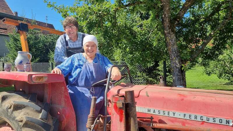 Zwei Personen sitzen auf einem roten Massey-Ferguson-Traktor. Sie l&auml;cheln und tragen l&auml;ndliche Kleidung. Im Hintergrund sind B&auml;ume und ein Haus zu sehen., &copy; SBEntertainment GmbH & Co. KG