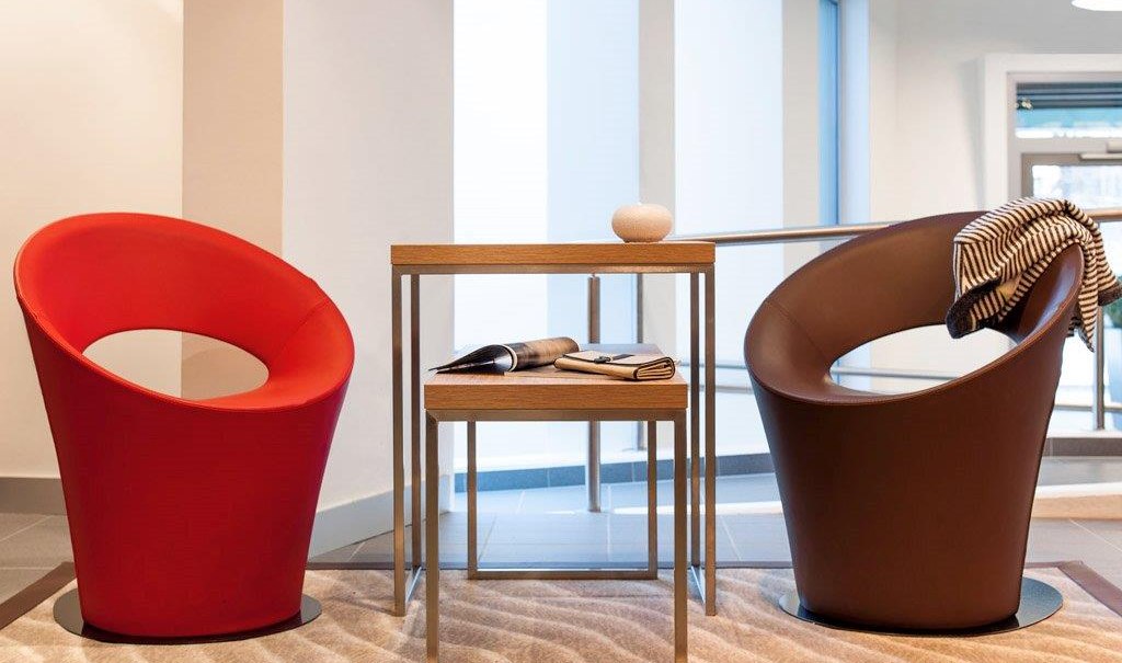 Zwei moderne Sessel, einer rot, einer braun, stehen in einer Lobby. Dazwischen ein kleiner Tisch mit Zeitschriften und Dekoration., © TOMAS