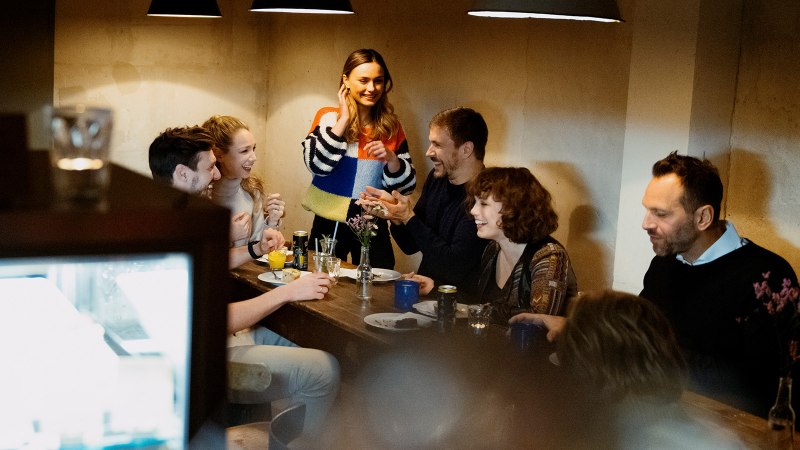 Eine Gruppe von Menschen sitzt in einem Café um einen Tisch und lacht. Eine Frau steht und spricht mit den anderen. Die Atmosphäre ist gemütlich., © Stuttgart Marketing GmbH, Alwin Maigler Eine Gruppe von Menschen sitzt in einem Café um einen Tisch und lacht. Eine Frau steht und spricht mit den anderen. Die Atmosphäre ist gemütlich., © Stuttgart Marketing GmbH, Alwin Maigler