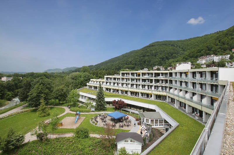 Modernes Geb&auml;ude mit Terrassen in gr&uuml;ner Umgebung und H&uuml;geln im Hintergrund. Ein Spielplatz und Sitzbereich sind im Vordergrund sichtbar.