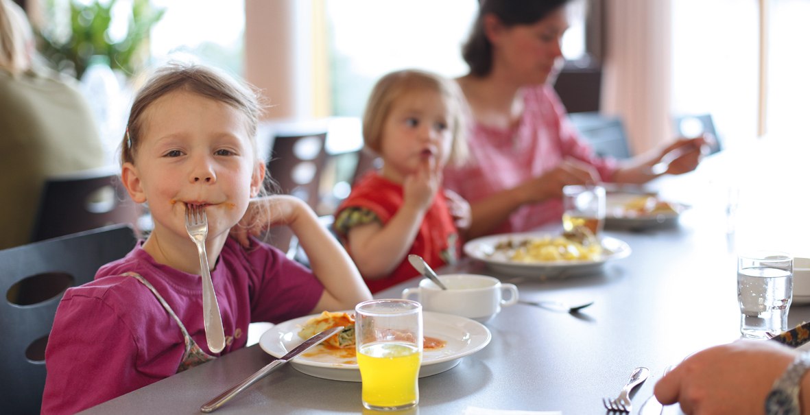Zwei Kinder und eine Frau sitzen an einem Tisch in einem Speisesaal und essen. Ein Kind hält eine Gabel im Mund. Auf dem Tisch stehen Teller und Gläser., © DJH Landesverband Baden-Württemberg e.V.
